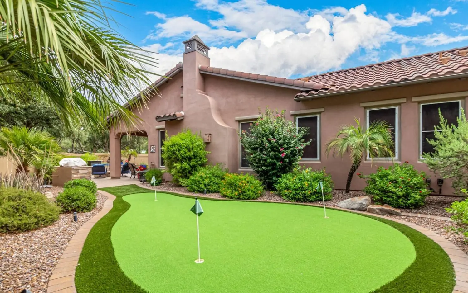 A charming backyard features a small putting green surrounded by neatly trimmed bushes and palm trees. The stucco house with its red-tiled roof overlooks the scene, while a grill rests in one corner. Under partly cloudy skies, it's an ideal space for enjoying backyard putting greens.