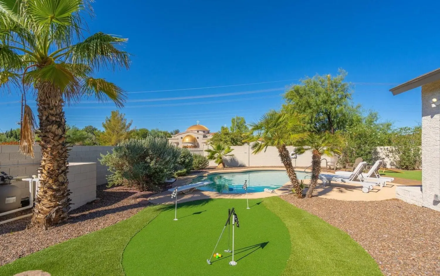 The backyard features a small putting green complete with two golf flags and lush landscaping. A sparkling swimming pool with lounge chairs invites relaxation, nestled among palm trees and vibrant plants under a clear blue sky—truly the perfect oasis for putting greens enthusiasts.