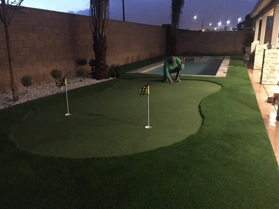 A person is setting up their backyard putting green at dusk, surrounded by artificial turf and marked with two flagged golf holes. Adjacent to the area, a narrow swimming pool shimmers in the evening light, while a brick wall defines the serene perimeter.