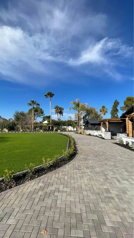 A sunlit brick pathway, reminiscent of expert paver installation, curves alongside a well-manicured lawn with palm trees and bushes. The sky is clear with wispy clouds, while a house with a wooden facade peeks from the right and mountains rise majestically in the background.