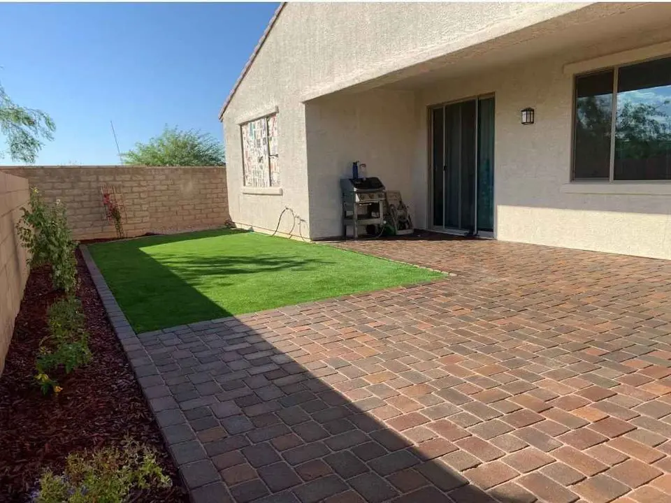 A backyard showcases a patio with expertly crafted brown paver installations and a small patch of green artificial grass. Near the sliding glass door of a light-colored house, a barbecue grill sits, complemented by a stone wall adorned with lush plants in the background.