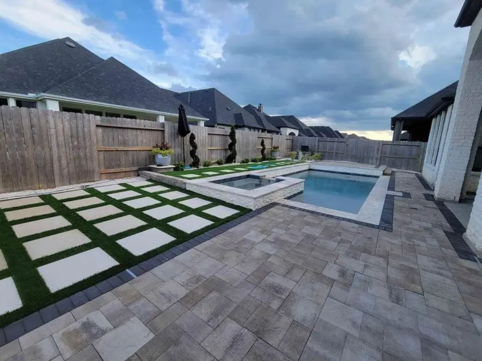 Modern backyard featuring a swimming pool and hot tub. The patio boasts beige stone tiles paired with expertly done paver installation, forming a neat grid of grass and stone squares. A wooden fence encloses the area, with neighboring houses visible under a partly cloudy sky.