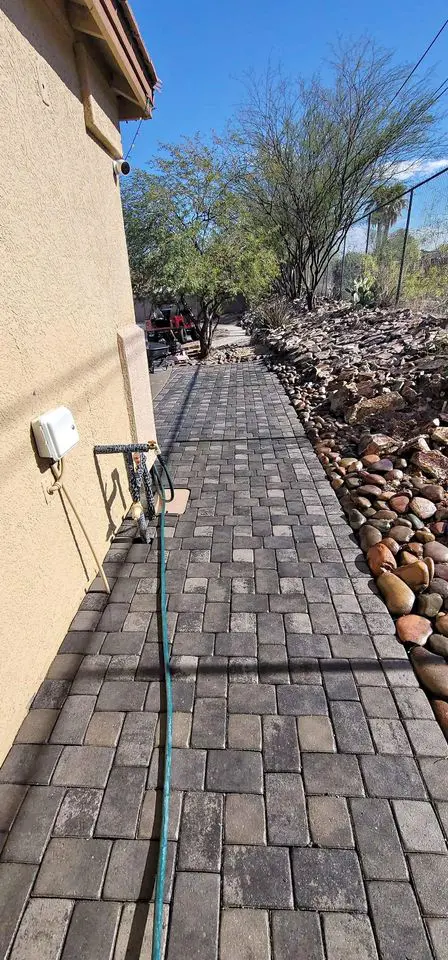 A paved path, the result of meticulous paver installation with rectangular stones, runs alongside a house. Nearby, a green hose is coiled on the ground. To the right, a rocky landscape features desert plants against a backdrop of clear blue sky.