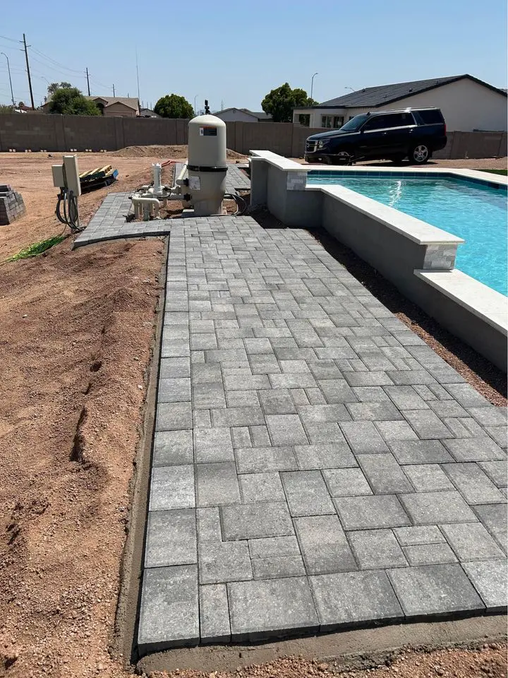 A newly completed paver installation features a gray path leading to a swimming pool, elegantly enclosed by a low wall. Nearby, a black SUV rests on bare dirt landscaping, with houses silhouetted in the background under a clear sky.