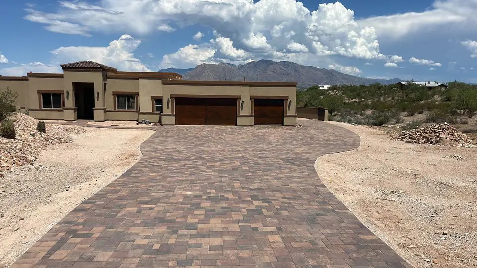 A modern, single-story house with a flat roof and beige exterior sits at the end of a long, expertly crafted paver installation. The garage boasts wooden doors, surrounded by desert vegetation under a partly cloudy sky with mountains in the background.