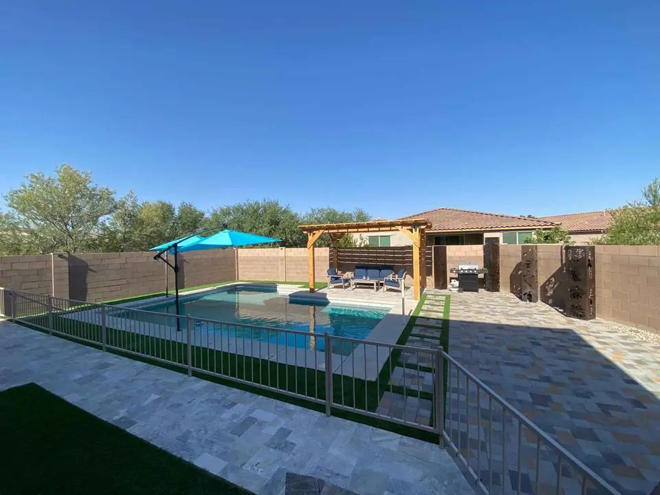 A backyard oasis features a sparkling pool enclosed by a fence. A seating area with blue cushions sits under a wooden pergola, while an outdoor kitchen boasts a grill. A turquoise umbrella provides shade over the pool, all set on beautifully crafted paver installation of colorful stones.