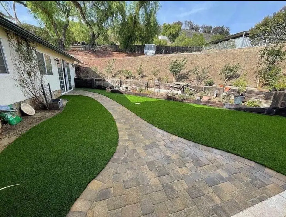 A backyard features a paved walkway, expertly designed with paver installation, curving through neatly manicured artificial grass. On the right, a slope hosts various potted plants and small trees. A white building is on the left, while tall trees border the background.