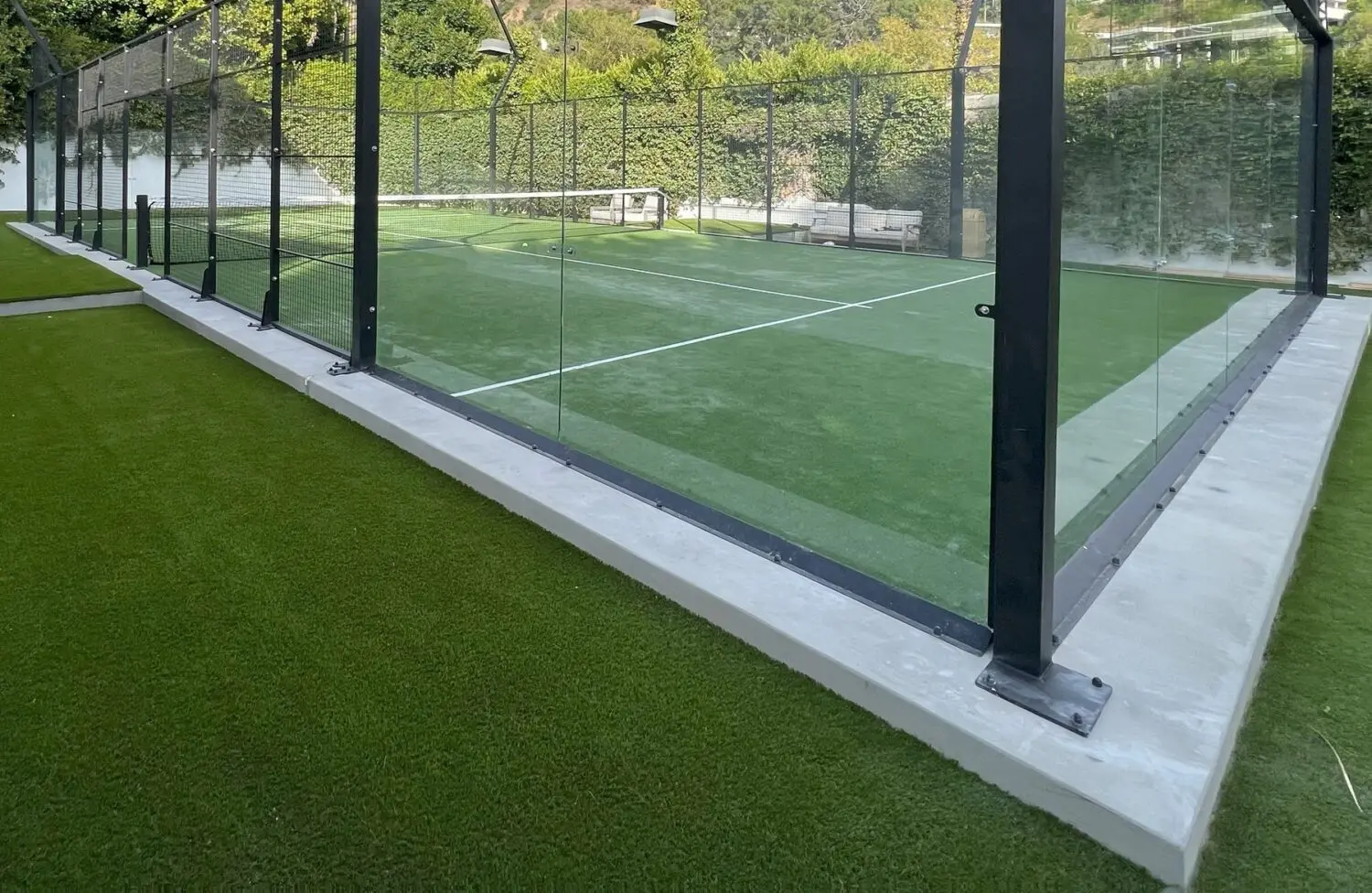 Gaze upon a padel court framed by glass walls, adorned with lush Vegas Artificial Turf. The vibrant green surface hosts a sturdy net, all set amidst flourishing greenery. Outdoor lighting enhances the scene, where a small bench quietly awaits in the background.