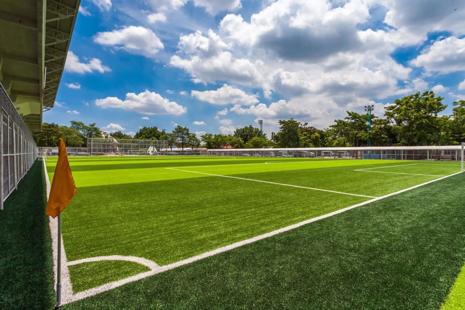 A well-maintained soccer field with lush, green Vegas Artificial Turf, an orange corner flag, and a clear blue sky dotted with clouds. Trees and a fence surround the field, while a small structure is visible on the left, completing this picturesque scene.