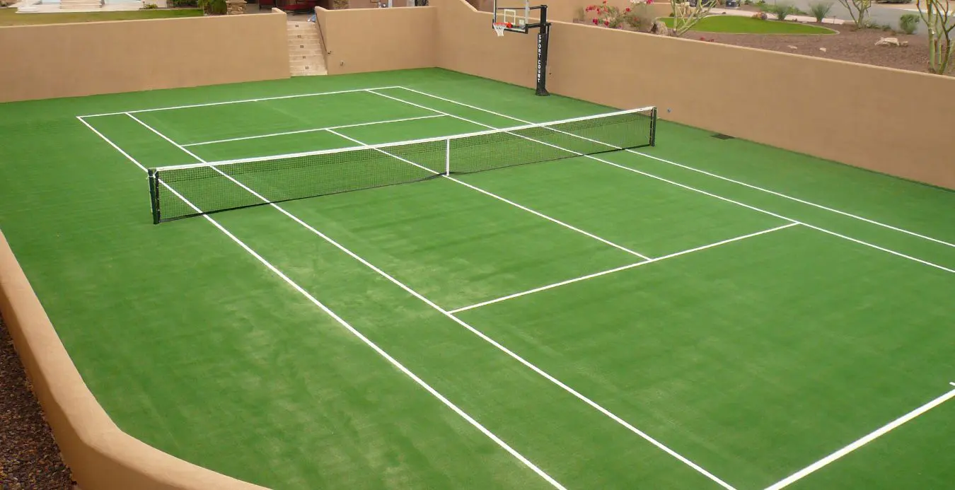 A vibrant pickleball court with Vegas Green artificial turf, marked by crisp white lines and a sturdy black net, sits in a walled outdoor area. A basketball hoop graces one end amidst desert landscaping, while steps ascend along one wall.