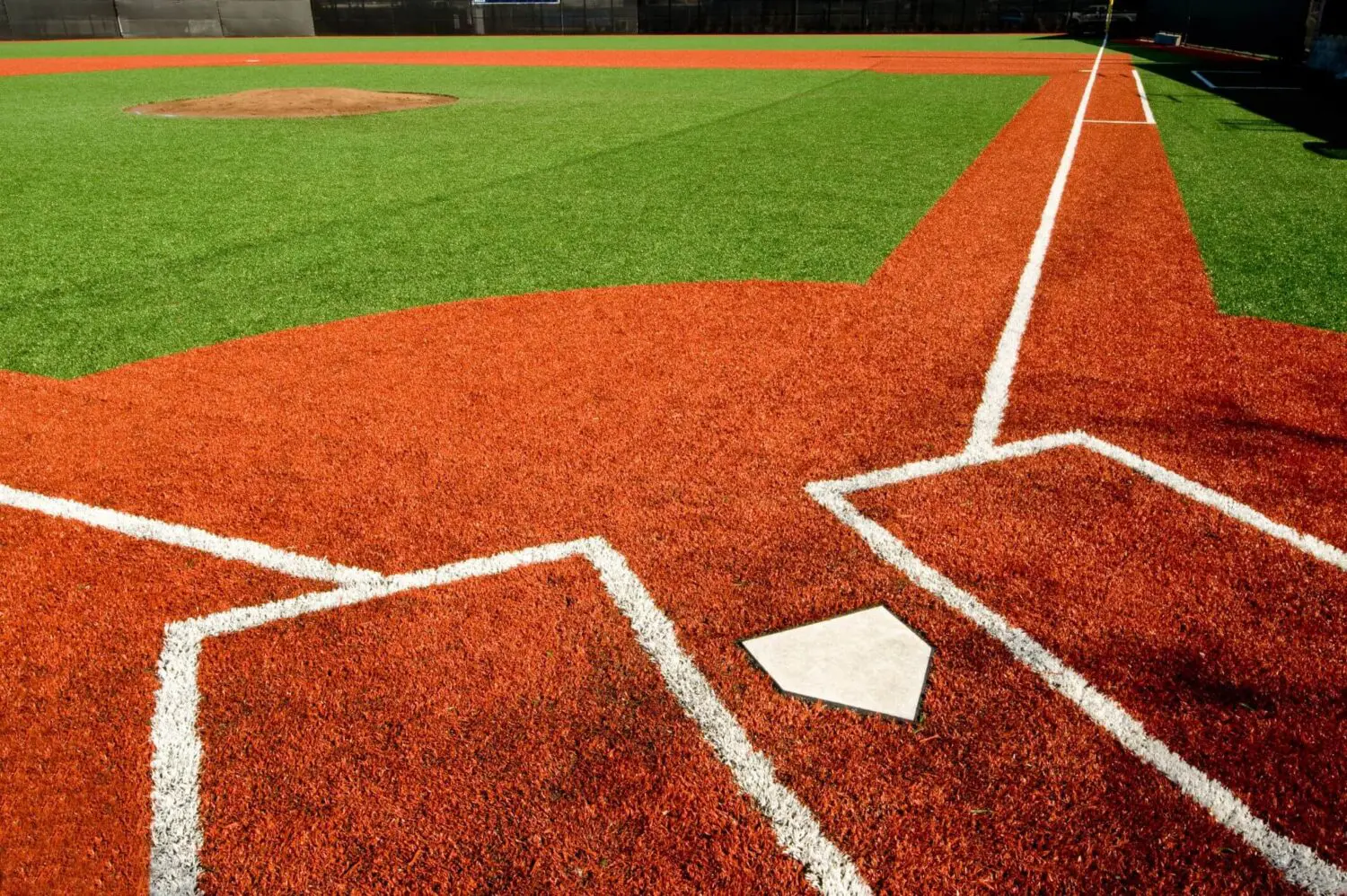 A baseball field with a focus on home plate and the infield area is vividly captured. The vibrant red clay contrasts sharply against lush Vegas artificial turf, while white lines crisply define the batter's box and baseline. The pitcher's mound is visible in the background.