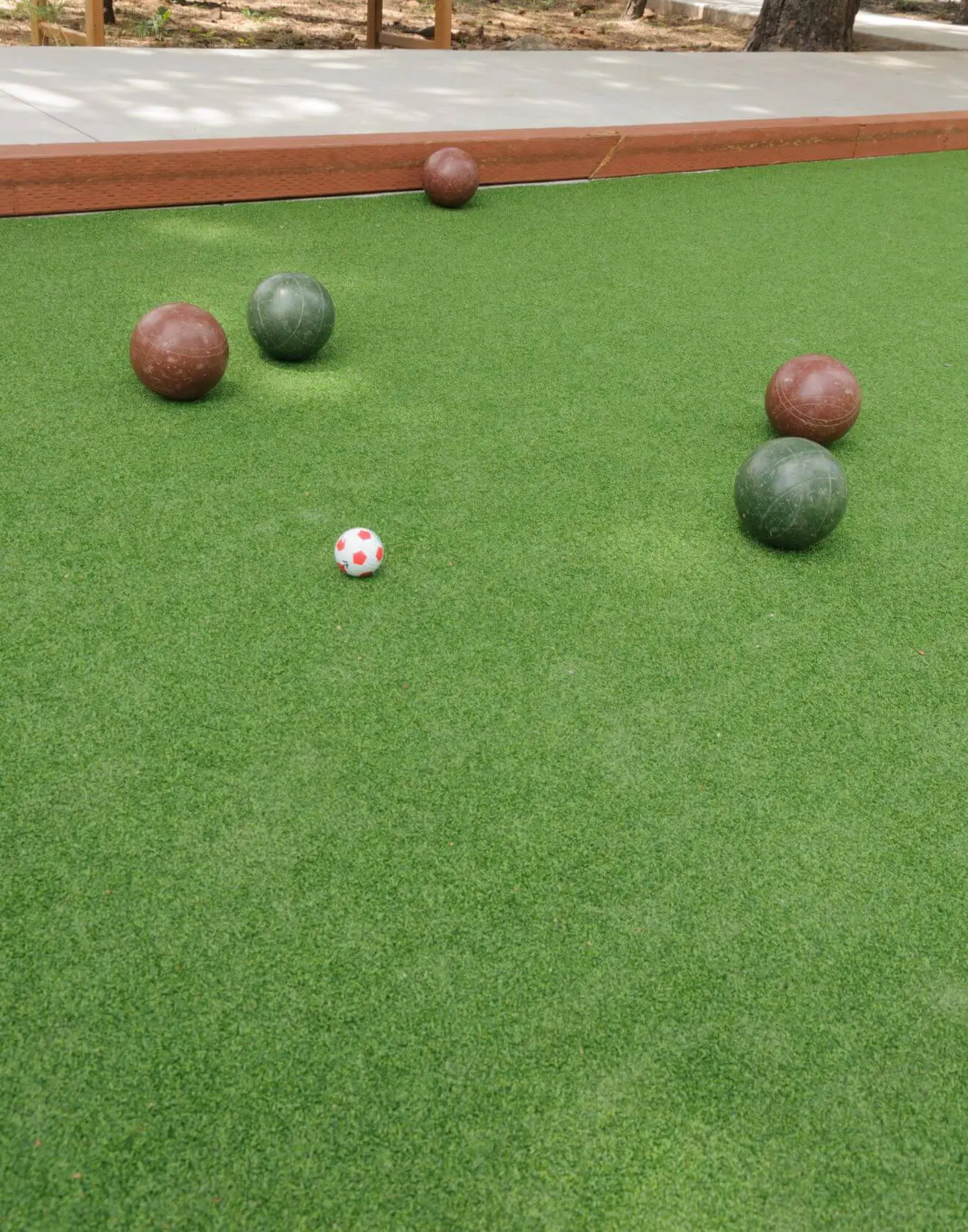 Bocce court with lush Vegas Artificial Turf, featuring five bocce balls—three green and two brown—scattered around. The small red and white pallino rests near the center, encircled by a sturdy wooden border.