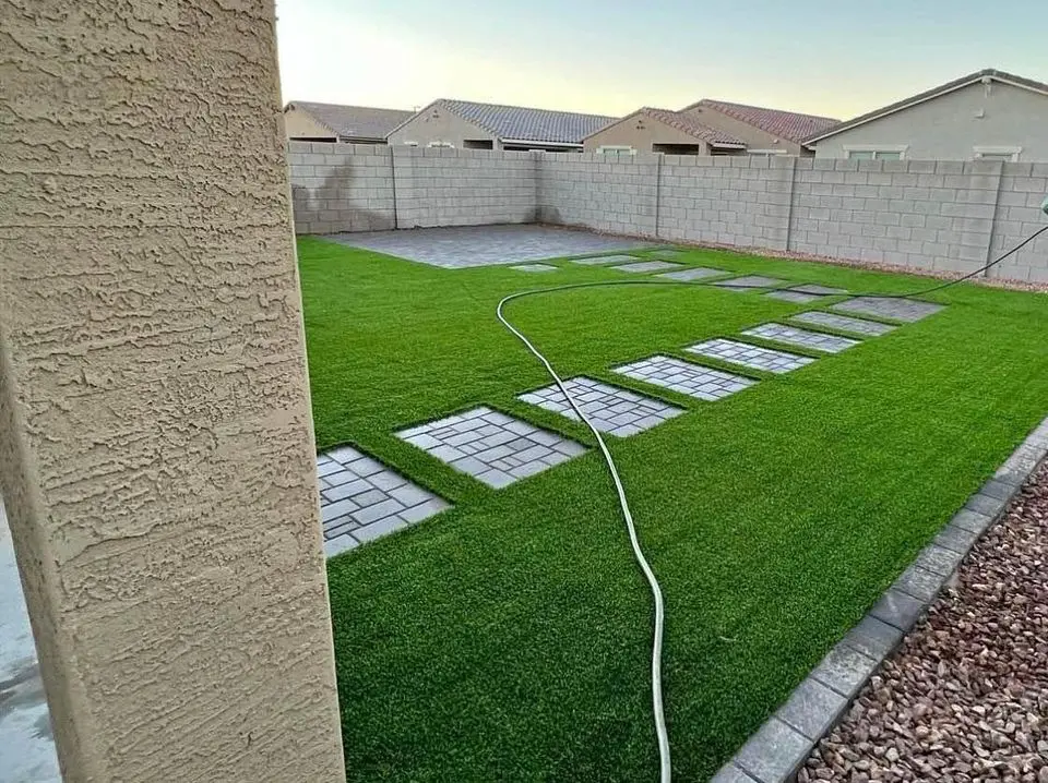 The backyard features a lush green grass lawn bordered by a concrete wall. A paved walkway with rectangular stones subtly enhances the Vegas aesthetic as it diagonally crosses the turf. A hose is coiled on the grass, while residential rooftops provide a serene backdrop.