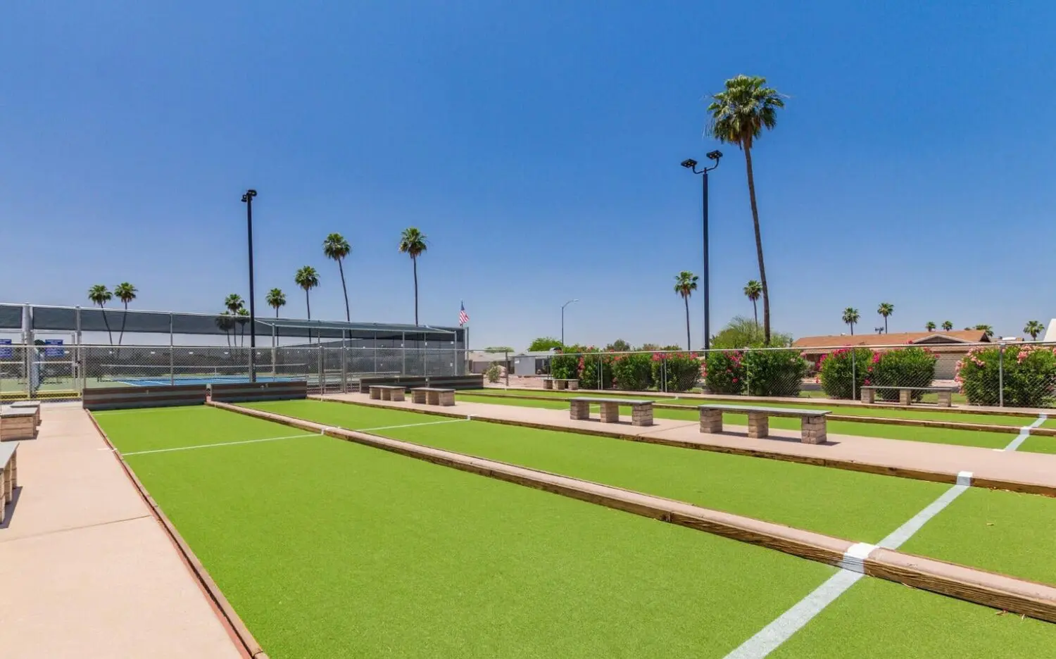 Outdoor bocce ball court featuring lush Vegas Artificial Turf under a clear blue sky. Surrounded by palm trees, the court boasts wooden boundaries and benches for seating. A netted area and a flag flutter in the background, enhancing this vibrant green oasis.