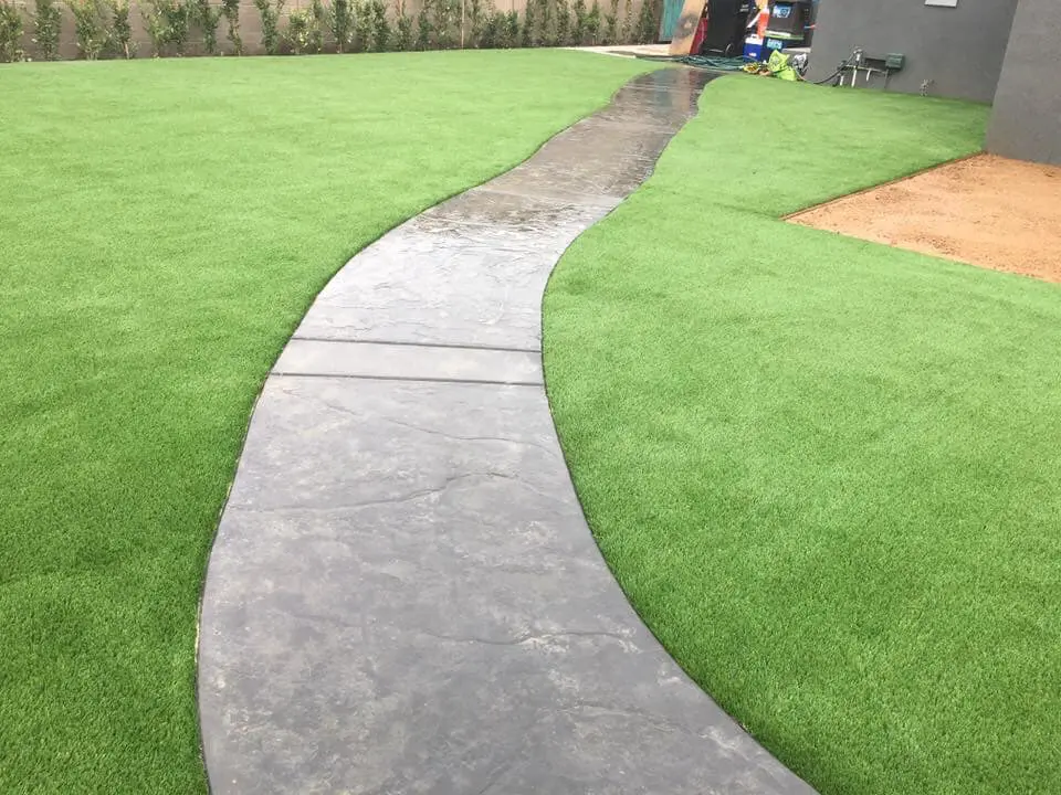 A winding, narrow concrete path cuts through a lush green lawn, leading towards a modern building in the background. Thanks to Vegas Artificial Turf and Green, the path is slightly wet, suggesting recent rain. Shrubs line the perimeter of this verdant landscape masterpiece.