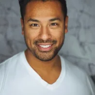 A person with short dark hair and a beard is smiling at the camera, exuding confidence like a seasoned paver installation expert. They are wearing a white V-neck shirt, while the background is a blurred, textured gray.