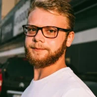 A man with a beard and glasses is looking at the camera, wearing a white t-shirt and standing outdoors. The blurred buildings and vehicles hint at an urban setting, possibly during sunset or sunrise. Perhaps he's contemplating his next paver installation project amidst this scenic backdrop.