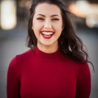 A smiling person with long dark hair wearing a red top stands outdoors, where the blurred background hints at an ongoing paver installation.