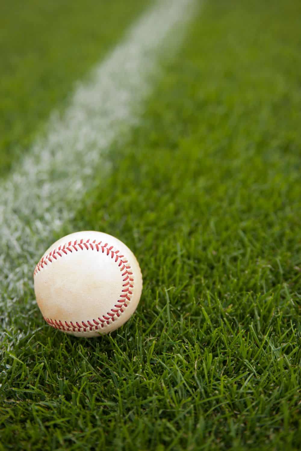 A baseball rests on a lush field of Vegas artificial turf near a white line. The grass is vibrant green, and the ball is slightly worn, showing red stitching.