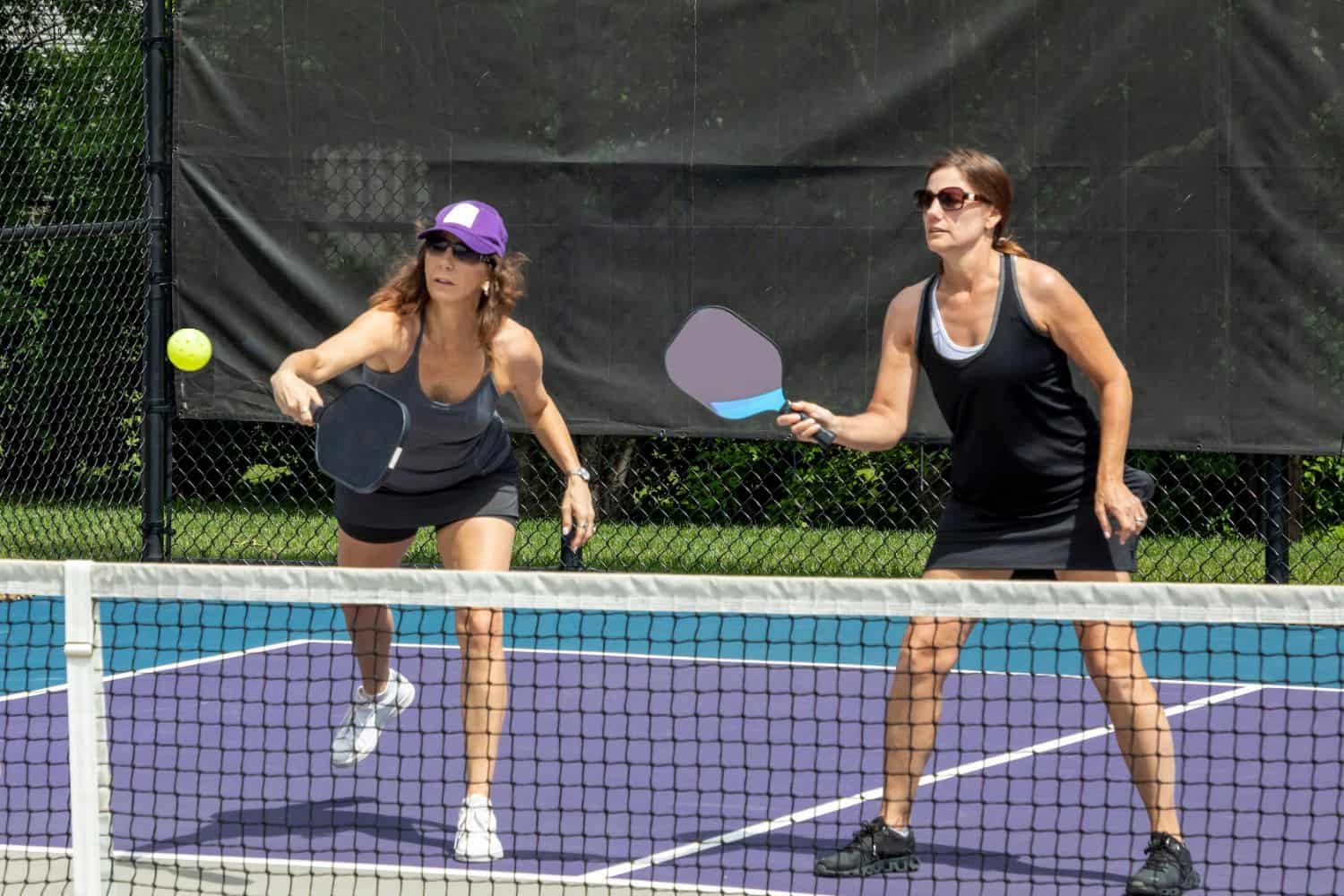 Two women enjoy pickleball on a court with vibrant purple flooring and a black net. One, in a purple cap and gray tank top, swings her paddle while the other, sporting sunglasses and a black tank top, eyes the yellow ball mid-air—a lively scene against the stunning Vegas green backdrop.