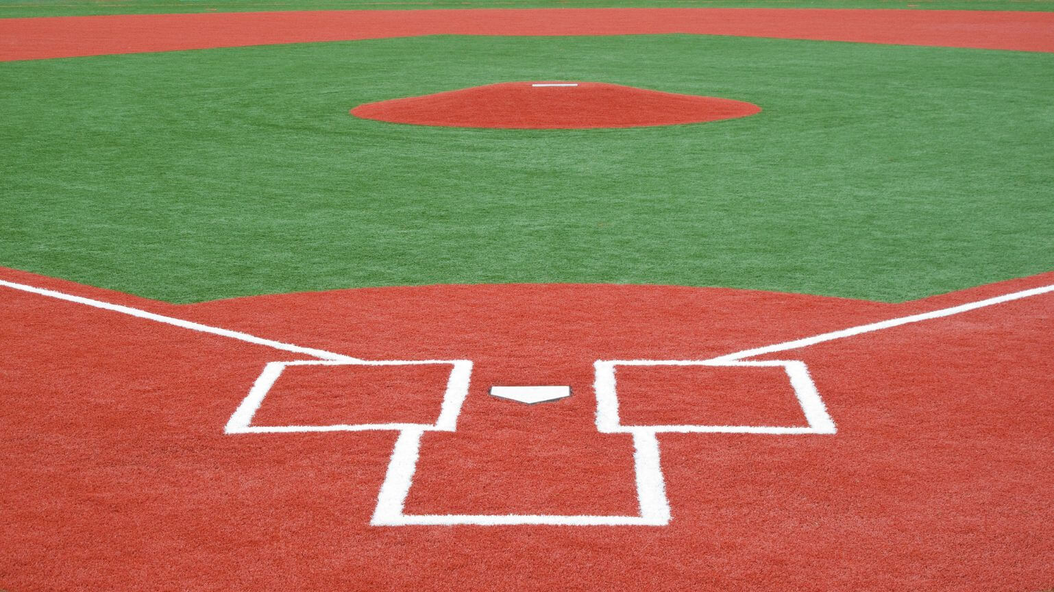 The view of a baseball field focuses on home plate and the pitcher's mound, where vibrant Vegas Artificial Turf contrasts with the clay-red base paths. White lines crisply mark the batter's boxes and base paths, enhancing the green expanse.